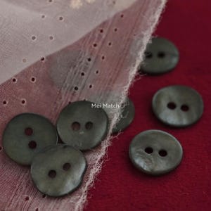 May include: A collection of round, dark olive green buttons with two central holes, scattered near a piece of sheer, light pink fabric with a perforated pattern. The text Mei Match is visible in the center of the image among the buttons.