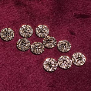 May include: Eleven gold-coloured buttons with a textured, floral design. Each button has four holes for sewing. The buttons are arranged on a deep burgundy fabric.