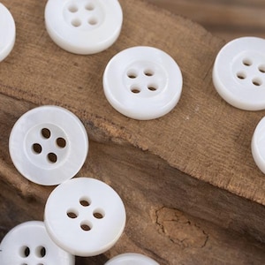 May include: A collection of white, round, four-hole buttons. The buttons are made of a smooth, shiny material and are arranged on a wooden surface.