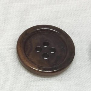 Peut inclure: Un bouton rond brun foncé avec une finition lisse et polie et un motif de grain de bois visible. Le bouton présente une zone centrale en retrait contenant quatre trous régulièrement espacés disposés en carré pour la fixation au tissu.