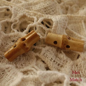May include: Two natural brown bamboo toggle buttons with holes for thread. The buttons are on a white crocheted fabric background.