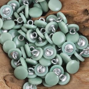May include: A collection of round, light green buttons with silver metal shanks. The buttons are scattered, showing their smooth, slightly domed surfaces. The buttons are a muted, pastel green color.