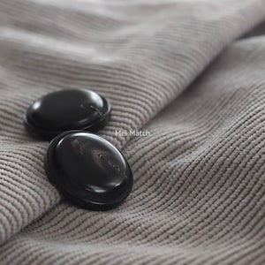 以下が含まれることがあります： 光沢のある滑らかな黒い丸いボタン2つが、ライトグレーの畝のあるコーデュロイ生地の上に置かれています。生地の質感のある表面、2つのボタンの間に小さな白い文字でMei Matchというテキストが印刷されています。