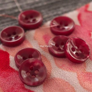 Pode incluir: Uma coleção de botões redondos vermelho-escuros, cada um com quatro furos no centro. Os botões têm um acabamento liso e brilhante e estão dispostos em grupo. O texto "Mei Match" está impresso em branco no centro da imagem.