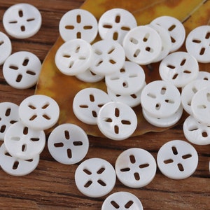 May include: A collection of white, round, shell buttons with four holes in a cross pattern. The buttons are scattered on a wooden surface and a leaf.