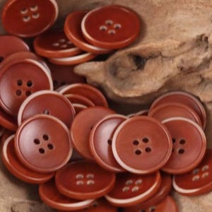 May include: A collection of round, reddish-brown buttons, each featuring four central holes and a distinct white rim. The buttons are stacked in a pile, showcasing their smooth, circular design and consistent color palette across the entire set.