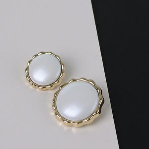 May include: Two circular buttons featuring a smooth, glossy white center inlay surrounded by a scalloped gold-toned metal rim. The buttons are arranged with one slightly smaller than the other, showcasing a reflective surface and a decorative edge.