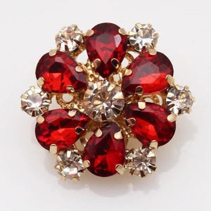 Puede incluir: Broche dorada con diseño floral. Presenta gemas rojas en forma de lágrima y gemas claras y redondas. Las gemas están engastadas en una base de metal dorado, creando un accesorio brillante y elegante.