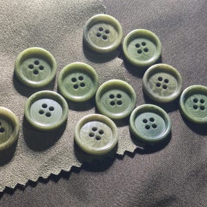 May include: A collection of ten round, sage green buttons, each featuring a four-hole center design and a subtle, swirling marbled texture. The buttons are arranged in a scattered pattern, showcasing their smooth, slightly raised circular edges.