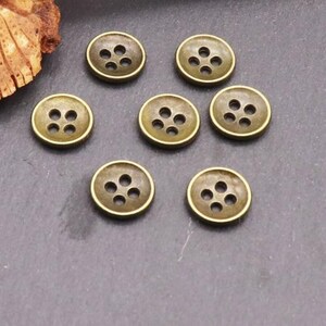 May include: Seven round buttons with a metallic bronze finish, each featuring four central holes arranged in a square pattern. The buttons have a raised outer rim and a smooth, slightly darkened surface texture, presented in a group.