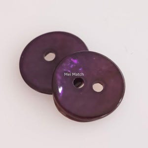 Puede incluir: Dos botones redondos de color morado oscuro con un acabado brillante y moteado. El botón frontal tiene dos agujeros para la fijación y el texto Mei Match impreso en blanco. El botón trasero es parcialmente visible y tiene un agujero central.