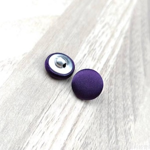 May include: Two round, purple fabric-covered buttons with a metal shank on a light wood surface. One button shows the metal shank, the other displays the solid purple fabric. Ideal for sewing projects.