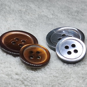 May include: Four buttons in varying sizes and colors are displayed. Two are a warm brown with four holes, and two are silver-toned with four holes. The buttons are round and have a glossy finish.