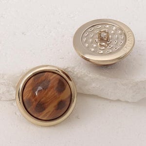 May include: Two round buttons with a gold-colored rim. The center of one button features a brown and tan animal print, while the other button's back is gold with a textured pattern and the text "Mei27323".