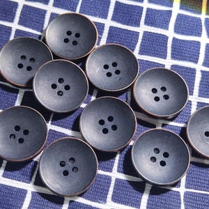 Puede incluir: Una colección de diez botones de color gris oscuro, cada uno con cuatro agujeros. Los botones tienen un brillo ligeramente metálico y un borde de color bronce. Están dispuestos sobre una tela a cuadros azul y blanca.