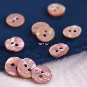 Peut inclure: Une collection de petits boutons ronds à deux trous avec une finition nacrée rose clair. Les boutons présentent une texture de surface lisse et légèrement irrégulière avec un éclat irisé subtil. Le texte Mei Match est imprimé au centre.