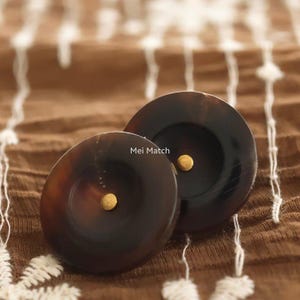 Puede incluir: Dos botones redondos de color marrón oscuro con un acabado liso y pulido y un centro cóncavo. Cada botón presenta un pequeño remache metálico de color dorado en el centro. El texto "Mei Match" está impreso en blanco entre los botones.