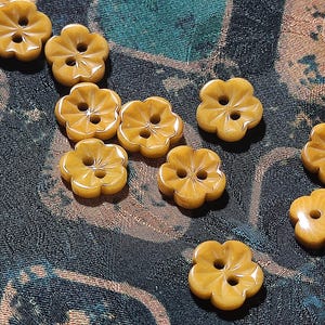 May include: A collection of nine mustard yellow buttons, each shaped like a five-petaled flower with a smooth, glossy finish and two central holes for attachment. The buttons are arranged in a scattered pattern, showcasing their uniform color and shape.