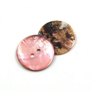 Może przedstawiać: Dwa okrągłe guziki. Jeden jest jasnoróżowy z dwoma otworami, a drugi brązowy z ciemnobrązowymi i beżowymi plamami. Oba guziki są na białym tle.