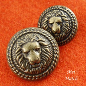 Op de afbeelding: Twee goudkleurige metalen knopen met een leeuwenkopontwerp. De knopen hebben een touwachtige rand rond de rand.
