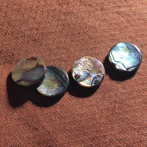 May include: Four circular buttons crafted from iridescent abalone shell, each displaying a unique swirling pattern of deep blues, greens, purples, and earthy brown tones with a polished, reflective surface that catches the light.
