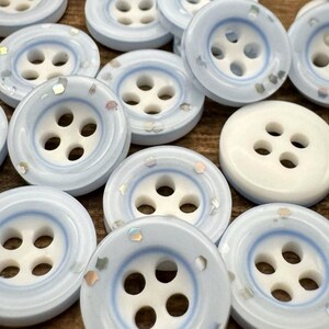 May include: Close-up of several light blue and white buttons with four holes. The buttons have a pearlescent finish and are scattered across a surface. One button is solid white. These buttons are ideal for sewing and craft projects.