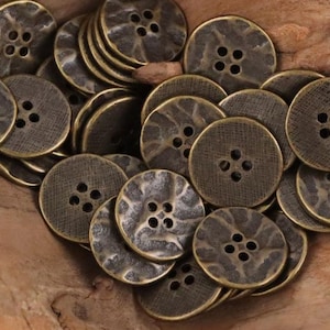 May include: A collection of antique brass-colored buttons with four holes each. The buttons have a textured surface and a slightly hammered appearance, suggesting a vintage or rustic aesthetic. The buttons are round and vary slightly in size.