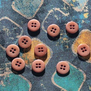 Könnte beinhalten: Eine Ansammlung von neun runden, hellbraunen Knöpfen, die jeweils vier in einem Quadrat angeordnete Löcher in der Mitte aufweisen. Die Knöpfe haben eine glatte, leicht glänzende Oberfläche und sind über den Rahmen verteilt, wobei sie eine konsistente Kreisform und eine einheitliche Farbe zeigen.