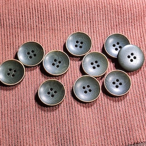 May include: A collection of ten round, four-hole buttons in a muted sage green color. Each button features a subtle, concentric wood-grain texture on its surface and a slightly raised, smooth rim. The buttons are arranged in a loose cluster.