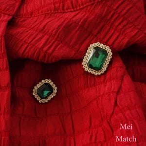 Puede incluir: Dos broches rectangulares de gemas verde esmeralda y doradas con un halo de pequeños cristales transparentes. Los broches están colocados sobre un fondo de tela roja texturizada. El texto "Mei Match" es visible en la esquina inferior derecha.