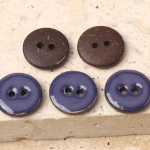 May include: Five buttons: two dark brown coconut shell buttons and three round, glossy, blue buttons. Each button has two holes for sewing. The buttons are arranged on a light-colored, textured surface.