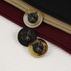 Puede incluir: Tres botones redondos con un diseño de conejo en relieve detallado. Los botones están hechos de un material marrón y blanco. Un botón es negro con un conejo dorado, y los otros dos son de color marrón claro con un conejo dorado.