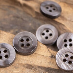May include: A collection of four-hole, dark gray, round buttons made of a smooth, shiny material. The buttons are arranged on a wooden surface.