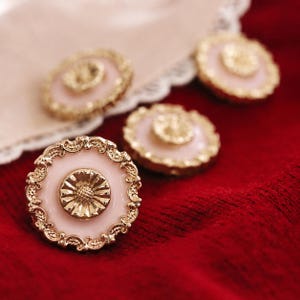 May include: Four decorative buttons with a floral design. The buttons have a light pink centre, a gold-coloured floral centre, and a gold-coloured filigree border. They are arranged on a red surface with a white lace trim.
