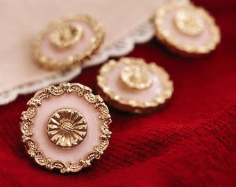 May include: Four decorative buttons with a floral design. The buttons have a light pink center, a gold-colored floral center, and a gold-colored filigree border. They are arranged on a red surface with a white lace trim.