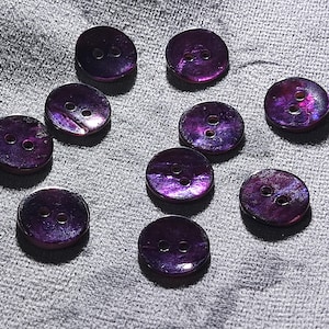 Peut inclure: Dix boutons ronds violets irisés avec deux trous chacun. Les boutons ont une finition brillante et sont dispersés sur un tissu gris. Les boutons sont d'un violet profond avec des reflets roses et bleus.