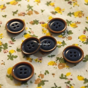 May include: A collection of navy blue buttons with four holes each, set against a floral patterned fabric. The buttons have a brown rim and are scattered across the surface. The text "Mei Match" is visible on some buttons.