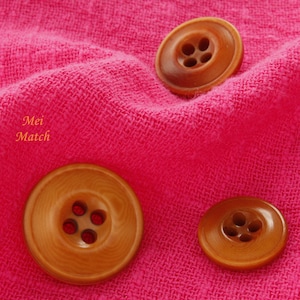 May include: Three wooden buttons with four holes each. The buttons are brown with a slightly reddish hue and are on a pink fabric background.