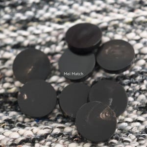 May include: A collection of eight circular, smooth, black buttons with a glossy finish. One button in the centre features the white text Mei Match printed on its surface. The buttons are arranged in a cluster, showcasing their uniform shape and dark colour.