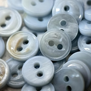May include: A collection of round, light blue buttons with a smooth, pearlescent finish. Each button features two central holes and a slightly raised outer rim. The surface of the buttons displays subtle variations in color and texture.