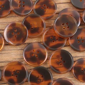 Puede incluir: Un conjunto de botones redondos con un patrón de carey. Cada botón tiene cuatro agujeros para coser y un acabado brillante. Los botones varían ligeramente en tonos marrones y ámbar.