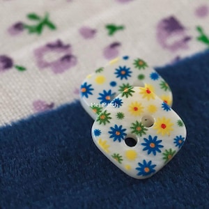 Puede incluir: Dos botones cuadrados blancos con un patrón repetitivo de pequeñas flores azules, amarillas y verdes. Cada botón tiene dos agujeros centrales para su fijación. El texto Mei Match está impreso en pequeñas letras blancas sobre los botones.
