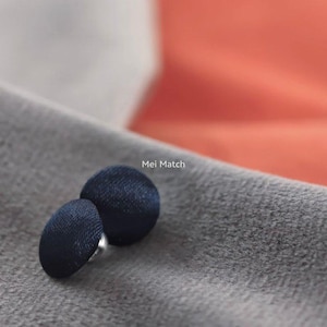 Puede incluir: Dos botones redondos cubiertos de tela azul marino oscuro con un acabado liso y satinado. Los botones están colocados juntos y el texto Mei Match está impreso en letras blancas cerca de los botones sobre la superficie de tela gris texturizada.