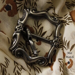 Peut inclure: Une boucle en métal argenté avec un design orné. La boucle présente un motif décoratif en spirale et un détail en forme de croix.
