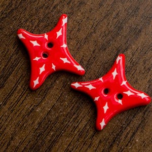 Puede incluir: Dos botones con forma de estrella de cuatro puntas en un color rojo vibrante. Cada botón presenta un acabado brillante y está decorado con un patrón repetitivo de pequeños iconos de estrellas blancas de cuatro puntas. Dos agujeros.