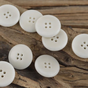 Op de afbeelding: Een verzameling van acht witte plastic knoppen met elk vier gaten. De knoppen zijn in een halve cirkel op een houten oppervlak gerangschikt.