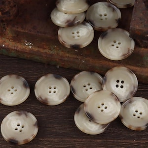 Puede incluir: Un conjunto de botones redondos de color crema con vetas marrones. Cada botón tiene cuatro agujeros para coser. Los botones están colocados sobre una superficie oscura y texturizada, ideal para un proyecto de costura.