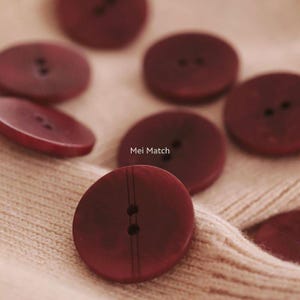 May include: A collection of burgundy-coloured, circular buttons, each with two holes, scattered on a cream-coloured fabric. The text "Mei Match" is visible on the fabric.