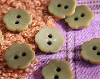 May include: Close-up of several olive-green flower-shaped buttons with two holes each. The buttons have a subtle orange trim and are scattered on a textured purple and brown surface. The text "Mei Match" is visible on one button.