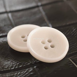 May include: Two round, off-white buttons with four holes each, featuring a smooth, slightly glossy finish. The text Mei Match is printed in small white letters on the surface of the front button, which is positioned slightly in front of the other.